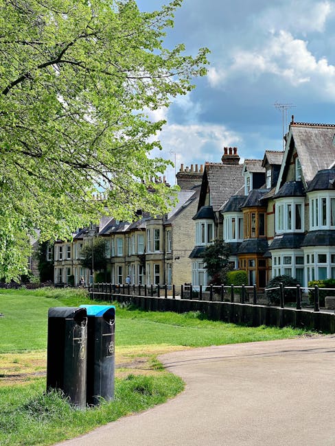 A row of traditional Victorian terraced houses with bay windows and pitched roofs is visible in the background, situated along a residential street under a partly cloudy sky. In the foreground, a grassy area with a large leafy tree provides partial shade, its branches extending across the scene. Two black cylindrical rubbish bins with blue lids are positioned at the edge of a pathway, which runs parallel to the row of houses. The bins appear to be for waste disposal, indicative of local or private rubbish collection services. Behind the bins, a low wooden fence separates the grassy area from the houses, with some shrubs and small trees adding greenery near the building facade. The overall scene captures a typical residential environment suitable for on-site waste management, with a focus on outdoor rubbish disposal in a quiet neighbourhood setting. This depiction aligns with private rubbish removal arrangements such as those managed by services like rubbishclearancestreatham.org.uk, illustrating an alternative to council-led rubbish collection for local residents.