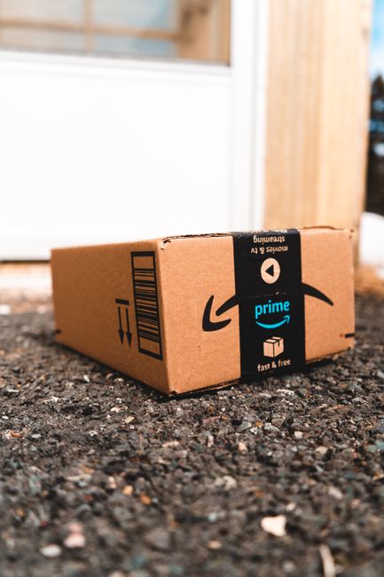 A small cardboard parcel, branded with Amazon Prime's black and blue tape, rests on an uneven, coarse gravel surface outdoors. The box appears to be recently delivered and is positioned close to a building with a white-framed window or door behind it. The surrounding environment includes a wooden fence panel on the right side of the image, suggesting a garden or backyard setting. The gravel ground is made up of dark, rough-textured stones with small scattered pebbles, and the box's materials are typical corrugated cardboard with a smooth finish on the printed surface. The Amazon Prime tape, prominently featured, is black with blue and white logos and text, securing the parcel with a clean, tight seal. Natural ambient lighting illuminates the scene, highlighting the subtle textures of the gravel and the matte finish of the cardboard. The image conveys an example of home delivery waste, which aligns with independent collection or private disposal practices often supported by rubbish removal services such as those on the Streatham Common drop-off guide, or in general waste management context.