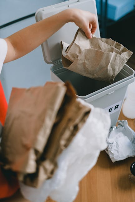A person places a crumpled brown paper bag into a grey rubbish bin with a black lid, which is positioned on a wooden surface. The person's arm is extended, and they are holding the paper bag by its edges, guiding it into the bin, while other paper waste is held in the foreground, partly obscuring the view. The bin appears to be situated indoors, possibly on a kitchen or utility room floor, with sufficient lighting illuminating the scene. The environment around the bin is tidy, with some additional crumpled paper visible beside it. This image highlights the process of waste disposal, aligned with private rubbish collection or on-site clearance services offered by companies such as rubbishclearancestreatham.org.uk.